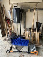 Photo of rakes, shovels, blue snow shovel, garage brooms, pair of wooden horses against wall in garage