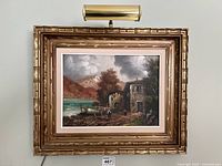 Framed oil painting showing lakeside buildings, water, figures, and mountains, lit by attached lamp above.