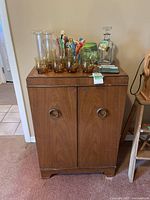 Front view of retro wooden bar cabinet with closed doors. Top surface holds glasses, ice bucket, decanter, drink stirrers, vase, and coasters.