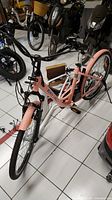 Peach Daymak Monte Carlo ebike frame with front and rear wheels, pedal, and one unmounted seat visible. Frame rests on white tiled floor with motorbikes in background.