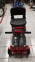 Front view of the red Daymak Boomer Buggy Mate electric mobility scooter showing black cushioned seat, handlebars with control panel, and two front wheels on white tiled floor.