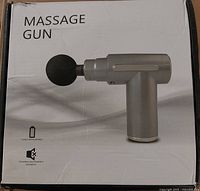 Front of the box showing image of the mini massage gun with black ball head attached. Highlights long-lasting battery and noiseless operation.