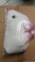 Photo of large white plush unicorn mother sealed in plastic bag resting on cardboard surface. Unicorn has pink snout, closed eye detail, and multicolor mane.