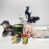 Photo displaying nine vintage ceramic animal figurines and planters including cat figurines, an elephant planter, a pig figurine marked Japan, a lamp planter with a crack, a sheep or lamb planter, and colorful toucan and parrot salt and pepper shakers.