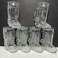 Front view of six clear cowboy boot shaped glasses arranged in two rows with detailed embossed floral and scroll designs.