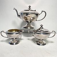 Photo showing three-piece silver tea set including teapot, sugar bowl, and creamer on white surface.