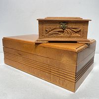 Both wood jewelry boxes, large light wood box with horizontal carved lines, smaller darker hand carved box with latch and foliage design
