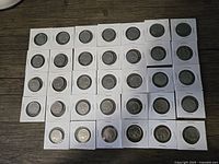 Overview of 34 Canadian 25-cent coins from 1973 RCMP Centennial series, each in individual white coin holders arranged in rows on wood surface.