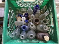 Top view of green plastic milk crate filled with 28 antique and vintage glass bottles of varying sizes, colors and shapes, some dusty and old, including clear, brown, and cobalt blue bottles.