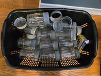 Photo showing a black plastic basket filled with approximately 20 vintage clear glass Crown canning jars, some with metal screw collars and sealing inserts. The jars appear dusty but intact.