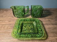 Complete set of three emerald green pressed glass items: creamer, sugar bowl, and square dish on wooden surface highlighting the color and beaded grape pattern.