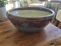 Side view of handmade pottery fruit bowl showing drip glaze in reddish-brown and bluish-gray on exterior, positioned on wood surface in natural light.