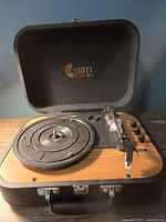 Open suitcase style vinyl record player showing the platter, tonearm, control knobs, and internal branding on lid.