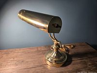 Front side view of the vintage brass bankers lamp showing the cylindrical shade, adjustable double curved arm, and round heavy base on a wooden surface.