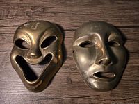 Two solid brass theatre masks shown from front, one comedy with smiling face, one tragedy with sad face.