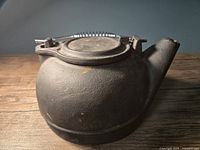 Front and top side view of the vintage cast iron kettle showing body, spout, lid, and handle lying flat on wood surface