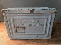 Front view of the metal ammunition box showing embossed 'CAL .30M1 AMMUNITION BOX' on faded white surface with wear and chipping.