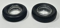 Two black rubber tire ashtrays with clear glass inserts placed side by side on a white surface.