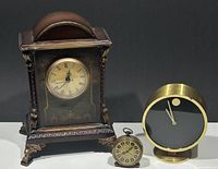 Three clocks arranged: a brown wooden mantel clock with gold embellishments, a small brass Lancel travel alarm clock, and a mid-century modern Howard Miller brass clock with minimalist black face.