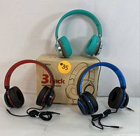 Front view of three wired over-ear headphones in red, blue, and teal placed around original packaging box.