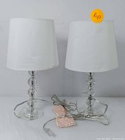 Pair of two table lamps with white tapered fabric shades and clear crystal-inspired bases, cords and plugs visible, on white surface.