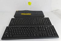 Three keyboards shown: one ergonomic wireless with wrist rest on top and two wired USB keyboards below it, all black with the ergonomic keyboard having yellow highlights.