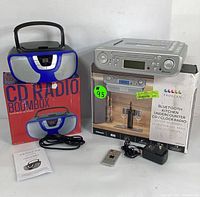 Lot showing both Proscan devices: the undercounter kitchen CD radio with digital clock and the blue CD boombox on their boxes, plus accessories including remote, power adapter, and manual.