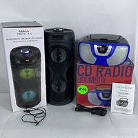 Front view of the Proscan Bluetooth LED speaker, the black cylindrical unit showing two speaker elements and multicolor LED lights, alongside the matching Proscan CD boombox in blue and black, box, power cables, and manual.