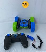 Front view of blue and green remote control stunt car with oversized wheels, remote control, battery, and USB charging cable arranged in front.