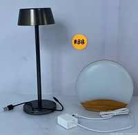 Both the black USB desk lamp and the round LED sunrise alarm clock with wooden base displayed side by side on a white surface, showing cables and power adapter.