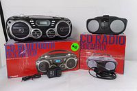 Photo of both Proscan audio units, their original red boxes, and power adapters, showing the Bluetooth CD boombox and the classic CD radio side by side.