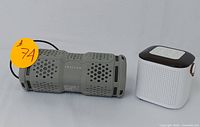 Two portable Bluetooth speakers shown side by side on a white surface; one is rugged olive green with perforated design and the other is white with a brown top and strap.