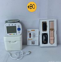 Digital blood pressure monitor with large screen and adjustable cuff, placed on a white base. Next to it is a silver-pink smart watch, a pink silicone strap, USB charging cable, and user manual.