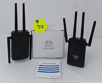 Two black Wi-Fi repeater units with four external antennas each, placed upright next to a white user manual with blue text. One repeater has 'WiFi Repeater' text, the other 'WiFi AP Repeater 1200M' text visible.