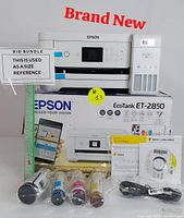 Brand new Epson EcoTank ET-2850 printer shown with original box, ink bottles, power cable, and installation materials. Blue protective tape on printer is visible.