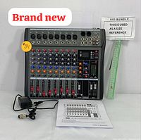 Photo of the front and top of the 8-channel audio mixer showing all input/output ports, control knobs, and sliders with power adapter and user manual placed in front. A size reference ruler and bidding sticker shown.