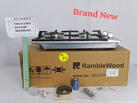RambleWood 2-burner gas cooktop displayed on top of its branded box alongside installation accessories and a battery for scale.