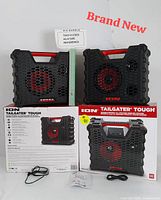 Front view of two ION Tailgater Tough speakers showing red-accented woofers behind honeycomb grills, with charging cables and packaging boxes in the background.