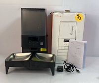 Full lot photo showing the FeelNeedy automatic pet feeder, dual stainless steel bowls set in black plastic base, original box, power adapter with USB cable, and printed user manual.