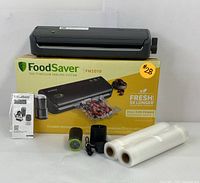 FoodSaver FM2010 vacuum sealer on top of original box with vacuum sealing rolls, vacuum accessory pump, power adapter, and instruction manual in front, all arranged on white surface.