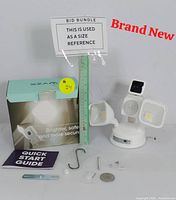 Photo showing the Wyze Smart Floodlight Camera, its box, Quick Start Guide, a metal hook, screws, and other mounting hardware next to a ruler for size reference