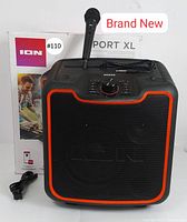 Front view of the speaker alongside its original box, wired microphone placed on top and power cable on the side. Clear brand and model labeling visible on box and speaker.