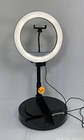 Front view of the functional illuminated LED ring light mounted on adjustable stand, showing the phone holder inside the ring and wired connection to the base with battery storage compartments.