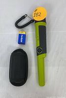 Handheld green and black metal detector with coiled tether, black fabric carrying holster, and blue 9V battery laid out on white surface.