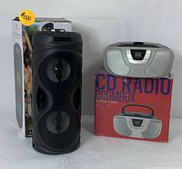 Photo showing black cylindrical Proscan Bluetooth party speaker next to silver Proscan CD radio boombox on original box.