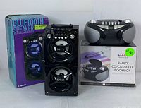 Photo showing Proscan portable Bluetooth speaker next to its box and Proscan CD/cassette radio boombox with its box in the background.