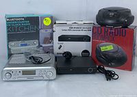 Three electronics items displayed together: Sylvania Bluetooth undercounter CD clock radio with packaging, HDMI upconvert DVD player with packaging and remote, and a portable CD radio boombox with box.