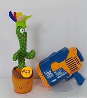 Dancing cactus plush toy with colorful fabric petals planted in a soft fabric pot alongside a blue and orange 132-hole bubble machine toy.