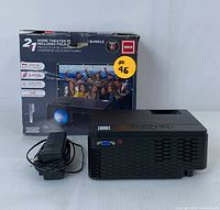 RCA home theater projector alongside its original packaging box and power adapter. Box indicates features like 2 HDMI inputs, 1080p support, and 50,000 hours LED lamp life.