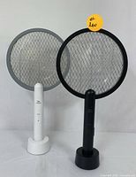 Two electric mosquito swatters, black and white, each with matching charging bases standing upright on a white surface against a white background.